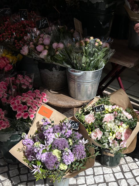 A display of fresh floral arrangements and bouquets at a flower market, featuring pink roses, purple alliums, and mixed pastel blossoms wrapped in brown paper and placed in metal buckets and on a wood
