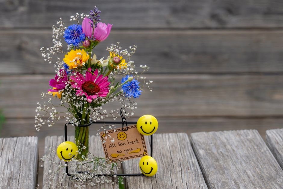 A colorful floral arrangement featuring a variety of fresh flowers including pink daisies, yellow calendulas, blue cornflowers, and purple bellflowers, all arranged in a clear glass vase. The bouquet