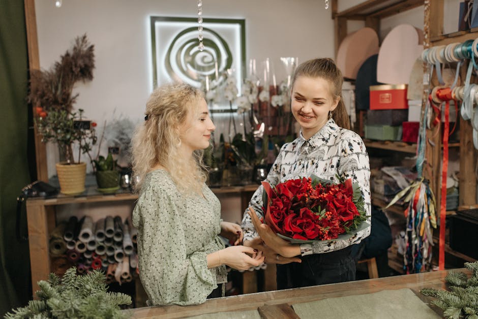 A floral arrangement featuring a bouquet of vibrant red roses, with some smaller red blossoms and greenery, is being handed over by a smiling young woman in a floral patterned shirt to a customer insi