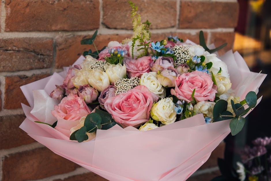A floral arrangement featuring a bouquet of fresh pink and cream roses, complemented by small white and blue flowers and sprigs of greenery, wrapped in pink tissue paper with a light pink ribbon. The